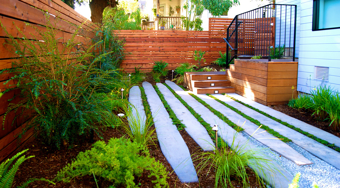 daytime view of backyard with outdoor lamps, paved walkway and plant beds.