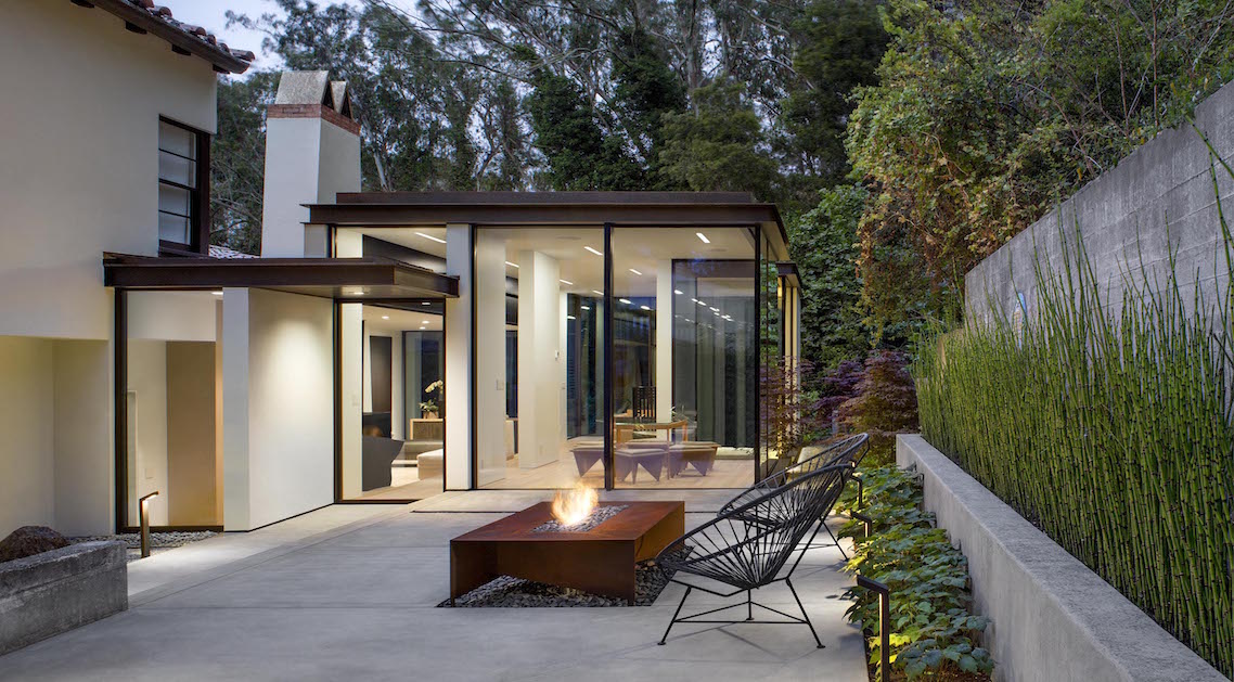 walled back yard with architectural landscaping including large metal firepit, seating area, concrete walls and planters. The home is visible in the background with large floor-to-ceiling glass walls.