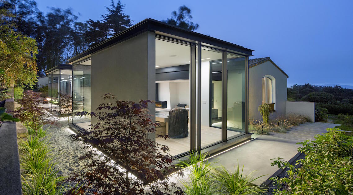 corner view of home at twilight, featuring recessed ground lighting and plants around the structure.