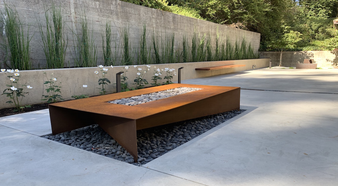 paved back yard with architectural landscaping including large metal firepit, concrete walls and built-in planters.