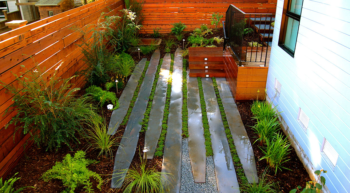 wide view of backyard with outdoor lamps, paved walkway and plant beds